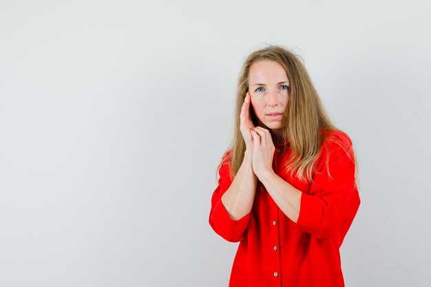 Signora bionda che tocca la pelle del viso sulla guancia in camicia rossa e sembra aggraziata.