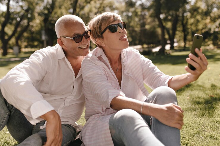 Signora bionda divertente in camicetta a righe e jeans che mostra la lingua e che fa foto con l'uomo sorridente in occhiali da sole e camicia all'aperto