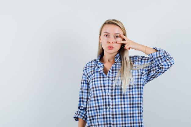 Signora bionda in camicia a quadri che mostra il segno di V, labbra imbronciate e sembra sicura