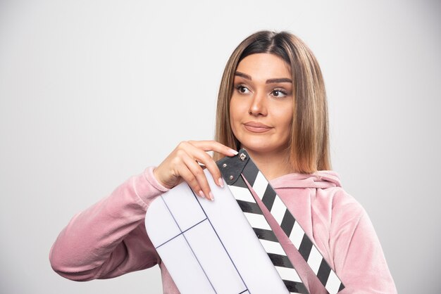 Signora bionda in felpa rosa che tiene un ciak vuoto e dà pose naturali