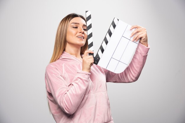 Signora bionda in felpa rosa che tiene un ciak vuoto e dà pose positive e divertenti.