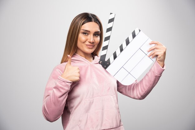 Signora bionda in felpa rosa che tiene un ciak vuoto e dà pose professionali.