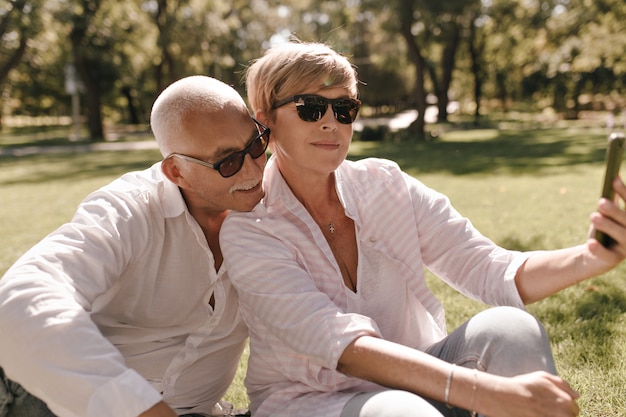 Signora bionda in occhiali da sole, camicetta elegante a righe e jeans. seduto sull'erba e fare selfie con uomo baffuto in camicia bianca all'aperto.