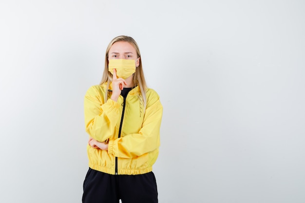 Signora bionda in tuta da ginnastica, maschera in piedi nella posa di pensiero e che sembra seria, vista frontale.