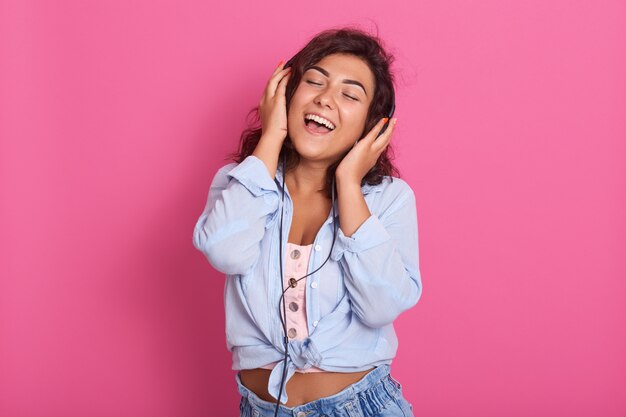 Signora bruna indossa camicia blu, jeans e top rosa, ascoltando musica in cuffia