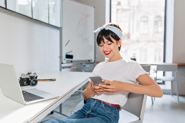 Signora castana allegra in camicia bianca e blue jeans che lavorano con il computer portatile in grande ufficio moderno