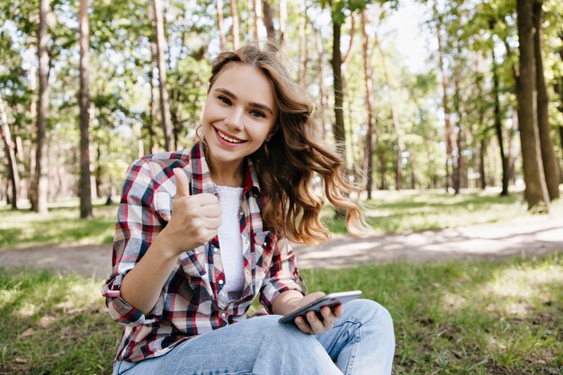 Signora caucasica soddisfatta che si siede per terra con il telefono in mano. Outdoor ritratto di gioconda ragazza bionda in abito casual in posa sul prato.