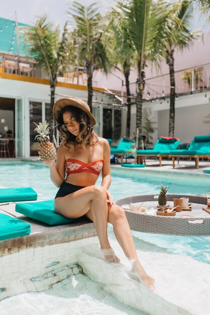 Signora caucasica sognante in cappello estivo alla moda agghiacciante in vacanza. Outdoor ritratto di incredibile donna riccia con ananas seduto accanto alla piscina.