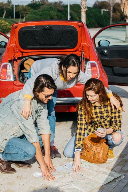 Signora che abbraccia la donna con lo zaino e smartphone vicino uomo e guardando la mappa vicino auto