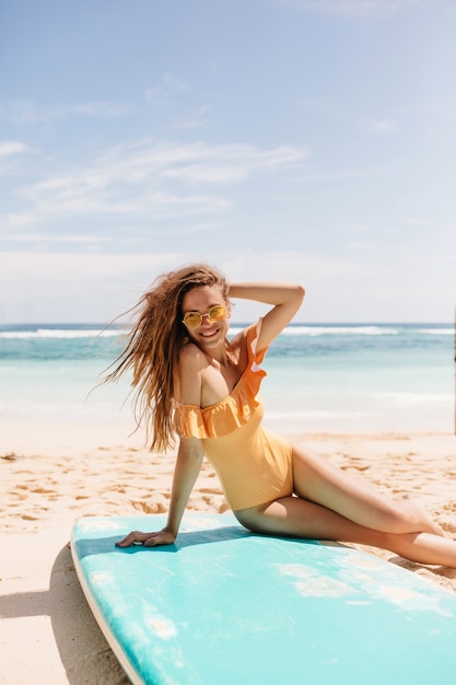 Signora che ride bruna posa in spiaggia dopo il surf. Magnifica ragazza in costume da bagno arancione seduto sulla sabbia e sorridente.