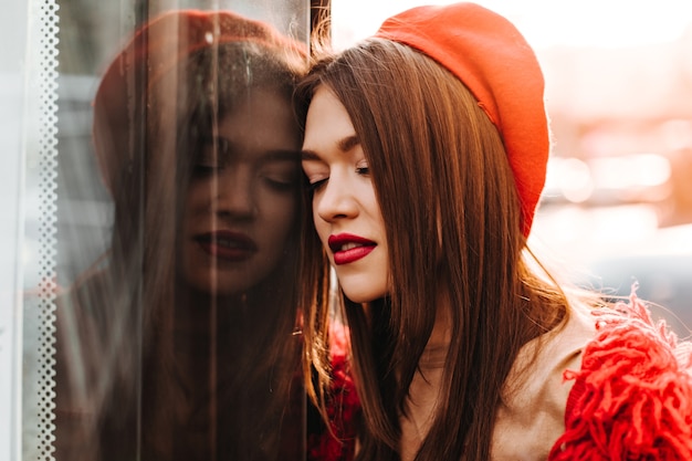 Signora con labbra rosse in berretto luminoso e maglione si è appoggiata alla finestra, in posa all'esterno.