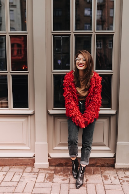 Signora con rossetto rosso in bicchieri, pantaloni in denim alla moda, cappotto luminoso sorridente e in posa fuori vicino alla finestra con cornice bianca.