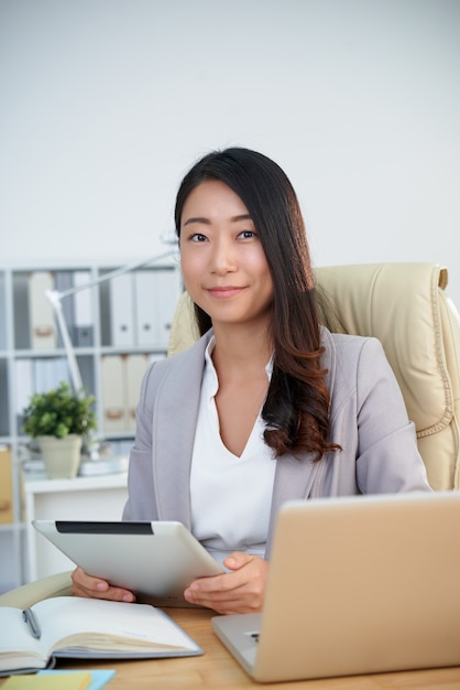 Signora coreana sorridente di affari che posa nell'ufficio con la compressa davanti al computer portatile