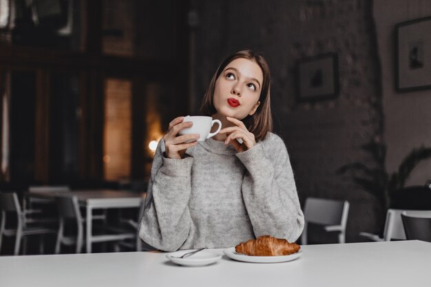 Signora dagli occhi marroni con rossetto rosso in posa minuziosamente con una tazza di tè. Donna in maglione grigio che si siede al tavolo con croissant.