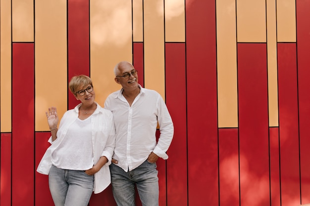 Signora dai capelli bionda in occhiali e camicetta leggera e fresca sorridente e in posa con il vecchio in camicia bianca e jeans su arancione e rosso.