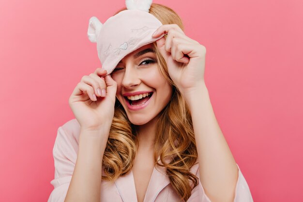 Signora dai capelli biondi estatica divertendosi la mattina presto. Affascinante ragazza europea in maschera per gli occhi divertente che ride sulla parete rosa.