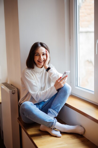 Signora dai capelli corti sorridente in blue jeans che si siedono sul davanzale e che scrivono messaggio sullo smartphone a casa