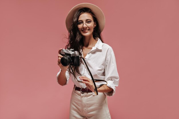 Signora dai capelli ondulati alla moda in cappello beige occhiali camicia bianca e pantaloni moderni sorridente e in posa con la fotocamera su sfondo rosa