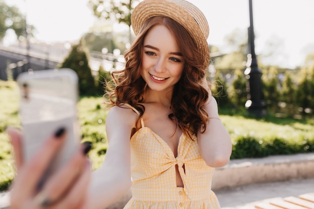 Signora dello zenzero Debonair in cappello che fa selfie nel parco verde. Foto all'aperto di affascinante donna europea con i capelli rossi che cattura maschera di se stessa sulla natura.