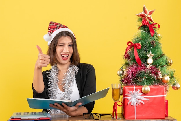 Signora di affari in vestito con cappello di babbo natale e decorazioni di capodanno che controlla il documento che fa il gesto giusto e si siede a un tavolo con un albero di natale su di esso in ufficio