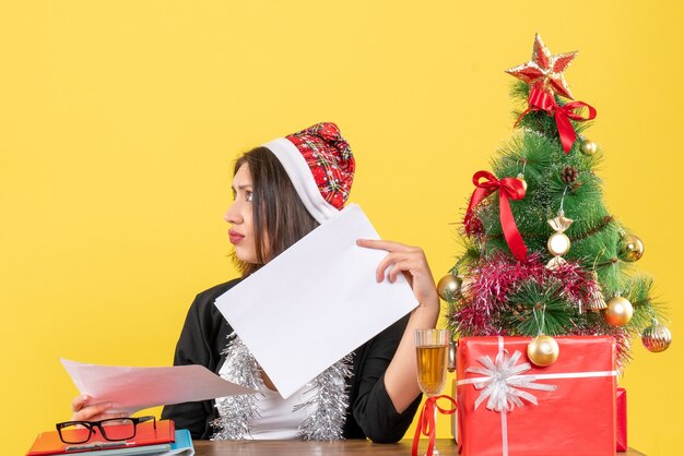 Signora di affari in vestito con cappello di babbo natale e decorazioni di capodanno sentirsi confusa e seduta a un tavolo con un albero di natale su di esso in ufficio