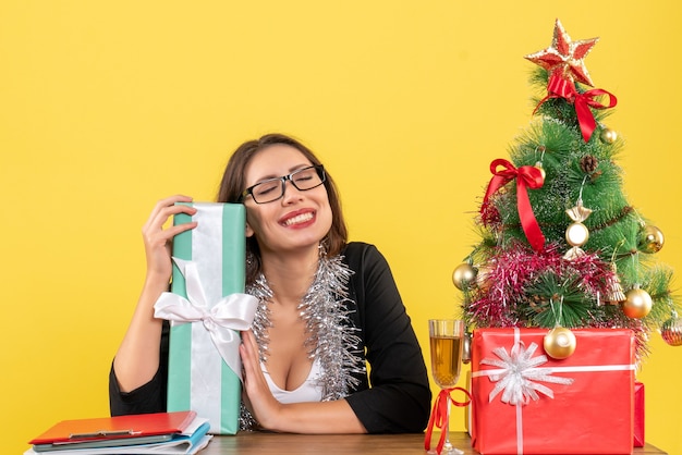 Signora di affari in vestito con gli occhiali che mostra il suo regalo che sogna qualcosa e che si siede a un tavolo con un albero di Natale su di esso in ufficio