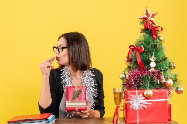 Signora di affari in vestito con gli occhiali che mostra il suo regalo chiudendo gli occhi e sognando e seduto a un tavolo con un albero di Natale su di esso in ufficio