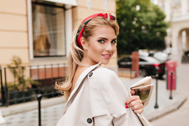 Signora entusiasta con nastro rosso in capelli biondi guardando sopra la spalla mentre cammina per strada