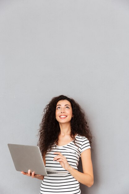 Signora felice con il messaggio di battitura a macchina dei capelli ricci o che comunica in Internet facendo uso del computer portatile d'argento che è isolato sopra la parete grigia