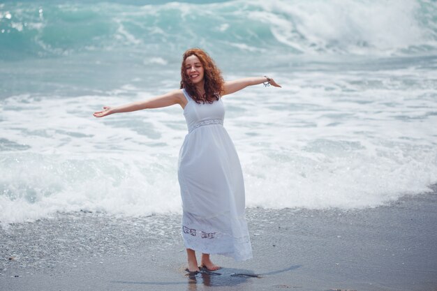 signora in abito bianco lungo in piedi le mani spalancate in spiaggia durante il giorno