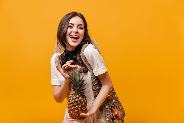 Signora in abito estivo di cotone ride e posa con ananas e borsa ecologica su sfondo arancione.