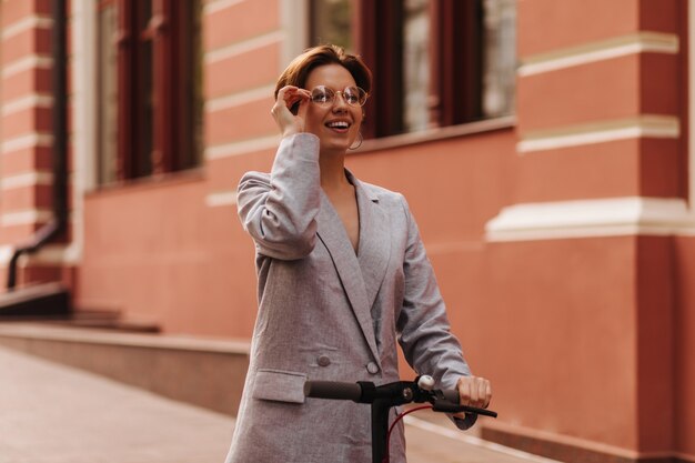 Signora in giacca grigia e occhiali da vista in sella a uno scooter elettronico. Felice donna eccitata in abito oversize ampiamente sorridente e godersi la città