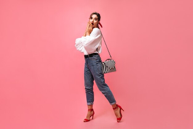 Signora in jeans e camicetta bianca sembra sorpresa su sfondo rosa. La giovane donna alla moda scioccata in scarpe rosse carine esamina la macchina fotografica con la borsa.