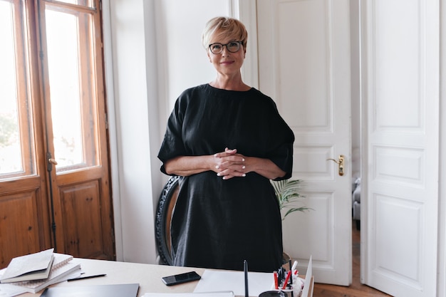 Signora in vestito nero e occhiali da vista esamina la macchina fotografica in ufficio