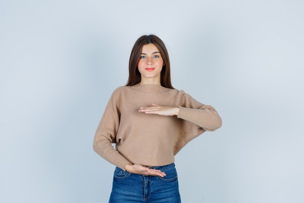 Signora meravigliosa che mostra segno di taglia in maglione, jeans e sembra allegra. vista frontale.