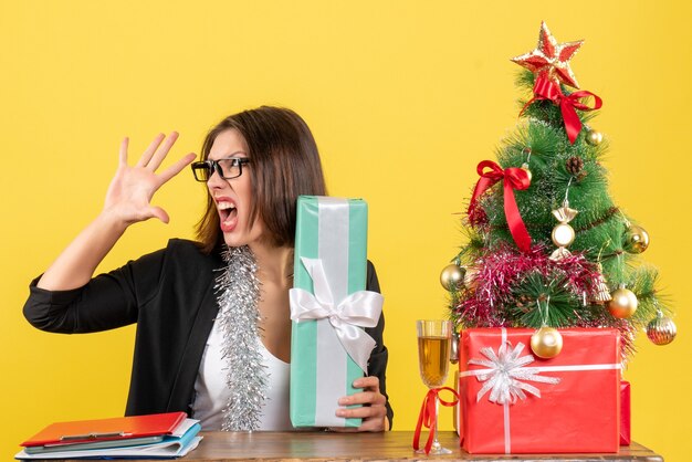 Signora nervosa arrabbiata di affari in vestito con gli occhiali che mostra il suo regalo e che si siede a un tavolo con un albero di Natale su di esso in ufficio