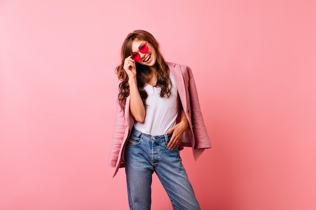 Signora ottimista dello zenzero in occhiali da sole luminosi che ride. Sorridente splendida ragazza in jeans in posa sul rosa.