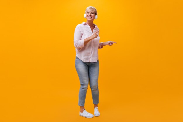 Signora positiva in jeans e camicia ballare e ascoltare musica in cuffia