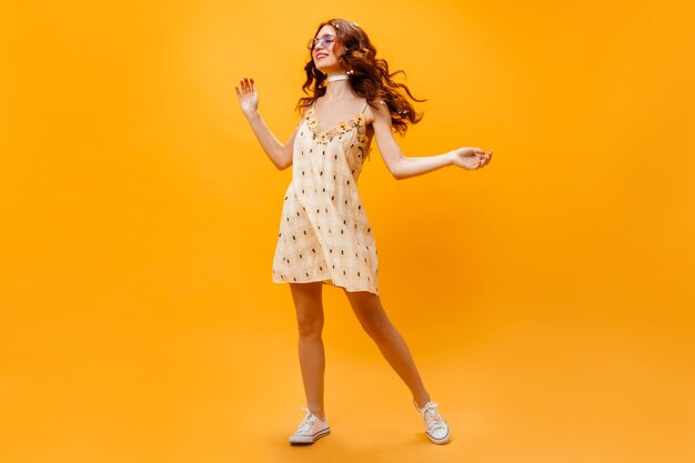 Signora riccia dai capelli lunghi in occhiali da sole e prendisole gialli che ballano su sfondo arancione