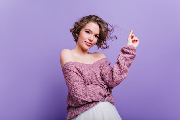 Signora sorpresa in eleganti scarpe rosa in piedi su una gamba e dito puntato. La foto dell'interno della ragazza carina riccia in maglione ha visto qualcosa di interessante.