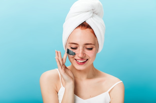 Signora sorridente con l'asciugamano sulla testa che applica la maschera per il viso. Studio shot di gioiosa ragazza caucasica facendo cure termali.