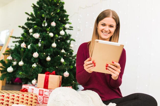 Signora sorridente con tablet vicino scatole regalo e albero di Natale