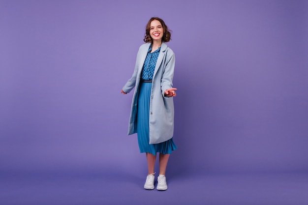 signora sorridente in elegante cappotto blu. Ritratto dell'interno della ragazza dai capelli corti che ride in scarpe bianche isolate sulla parete viola.