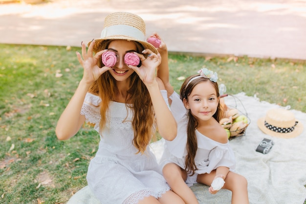 Signora sorridente in vestito bianco che tiene pan di zenzero rosa come occhiali, seduto su una coperta con la figlia. Bambina graziosa con il nastro che posa accanto alla madre scherzosa durante il picnic.