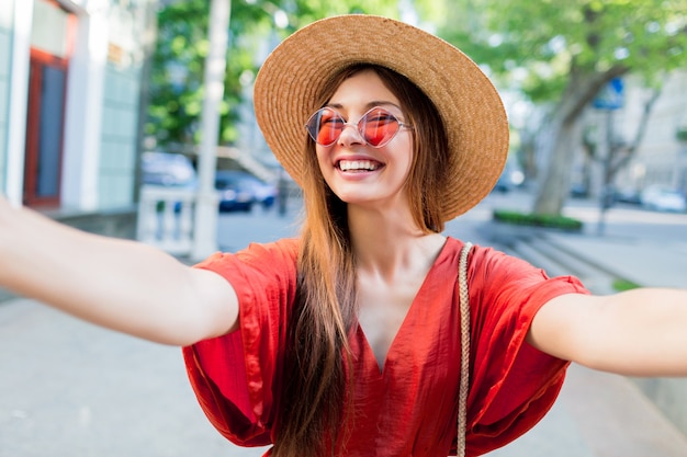 Signora sveglia in cappello alla moda che fa selfie mentre camminando all'aperto nei fine settimana estivi