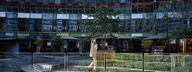 Silhouette di giovane donna d'affari in abito beige che cammina nel centro della città in posa vicino a edifici per uffici