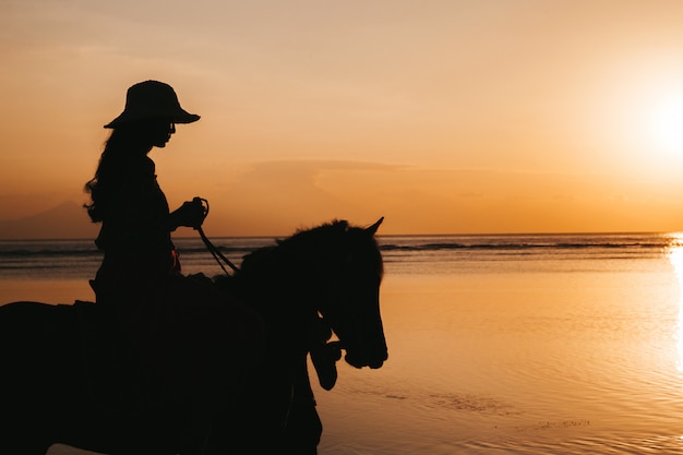 Siluetta della giovane donna che monta a cavallo sulla spiaggia durante il tramonto variopinto dorato vicino al mare