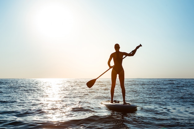 Siluetta di giovane bella donna che pratica il surfing nel mare all'alba.