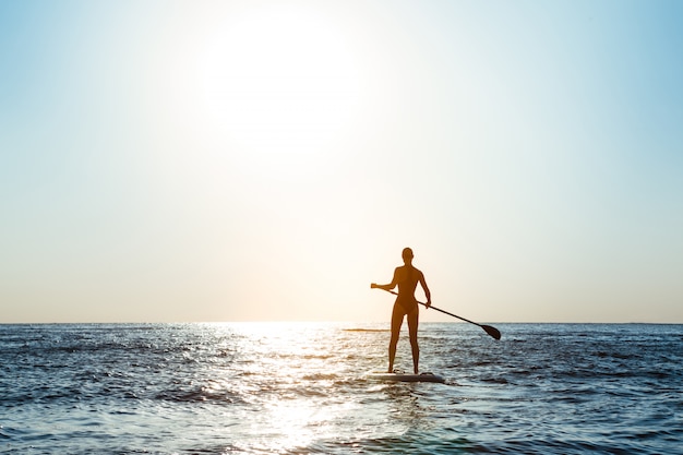 Siluetta di giovane bella donna che pratica il surfing nel mare all'alba.