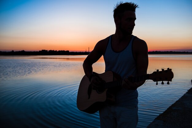 Siluetta di giovane uomo bello che gioca chitarra in riva al mare durante l'alba